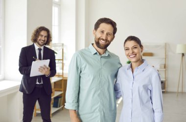 Arka planda erkek emlakçı olan genç ve mutlu bir çift yeni evde kameraya bakıyor. Profesyonel emlakçı yeni daireyi gösteriyor. Başarılı aile ev sahibi olmaya hazır..