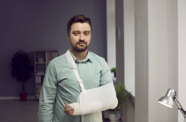 Kırık kollu genç adam, sakallı, kolunda yara bandı olan, evinde ya da işyerinde kameraya bakan sakallı bir adamın portresini takıyor. Tıbbi bakım, travmatoloji, fiziksel yaralanma