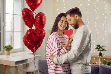 Elinde kalbi tutan esmer bir adam ve kadının portresi. Kapalı gözlerle sarılmış kırmızı folyo balonlarla. Aşık mutlu bir çift. Dikkat, şefkat ve sevgililer günü konsepti.