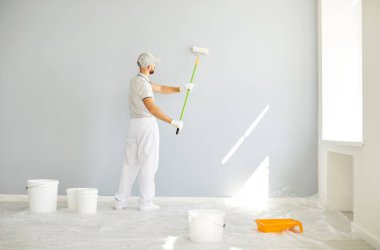 Beyaz üniformalı profesyonel bir erkek ressamın parlak geniş bir odada dikilip boya rulosuyla açık gri bir duvar boyaması, inşaat ve yenileme işleriyle uğraşması..