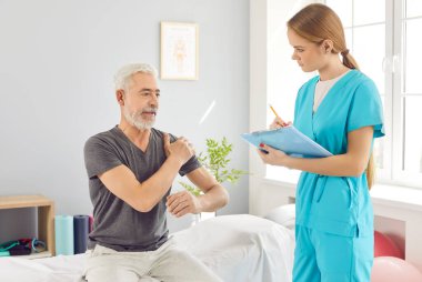 Modern rehabilitasyon kliniğinde kanepede oturan ve genç bir hemşireye ya da doktor fizyoterapiste omuz ağrısından yakınan olgun bir adam. Fizyoterapi ve fizik tedavi konsepti.