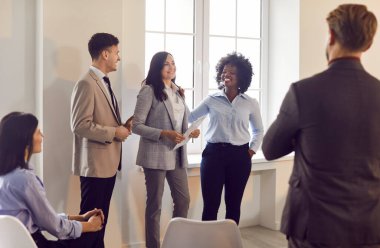 Çok kültürlü iş adamlarından oluşan neşeli bir grup bir arada duruyor, iş görüşmeleri veya eğitimleri sırasında bayan meslektaşlarını dinliyorlar. Gülümseyen başarılı erkek ve kadınlar şirket stratejisini tartışıyor.