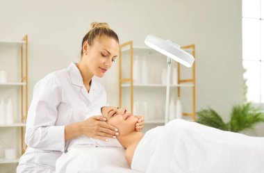 Bir masaj yatağında büyüteç altında uzanan ve sertifikalı bir güzellik uzmanının yaptığı yüz masajının keyfini çıkaran mutlu genç bir kadın. Cilt bakımı, güzellik, yüz bakımı kavramı