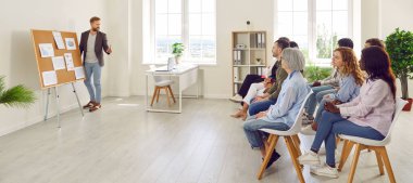 Çeşitli çalışanlar kurumsal iş eğitimi ya da seminerine katılıyor. Erkek işletme koçu, çok ırklı seyircilere sunum yapıyor. Modern hafif ofis içlerinde koltuklarda oturuyorlar.