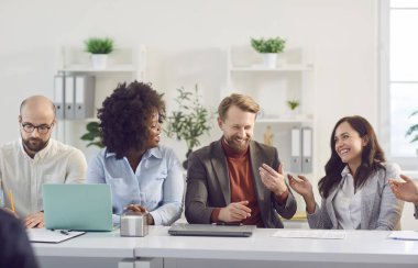 Mutlu gülümseyen çeşitli ofis arkadaşları toplantı masasında beyin fırtınası yapmak için toplanıyor, birlikte tartışıyorlar, çok ırklı iş arkadaşları ofiste iş birliği yaparak eğleniyorlar, takım çalışması, etkileşim.