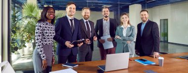 Çok ırklı iş adamları, kameraya bakıp içeride gülümseyen kadın ve erkekler. Şirket çalışanları ya da işyerindeki masada duran bir grup personel. Takım çalışması konsepti.