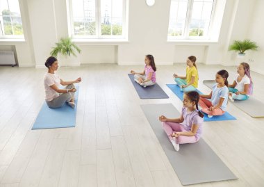 Yoga dersi alan bir grup çocuk. Çocuklar rahatlatıcı egzersizler yapıyor. Spor kıyafetli küçük kızlar nilüfer çiçekli minderlerin üzerinde oturup öğretmenleriyle meditasyon yapmayı öğreniyorlar.