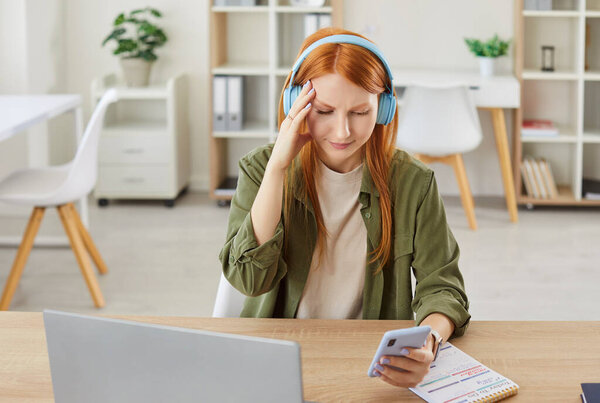 Young woman in wireless headphones at laptop holding phone, suffering from headache, migraine, touching face feeling stressed hearing bad news on cellphone, tired from too long talking on mobile phone