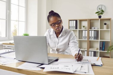 Ciddi, odaklanmış, güzel, gözlüklü, beyaz gömlekli Afro-Amerikalı bir kız. Masasında, geniş, rahat bir ofiste dizüstü bilgisayar monitörünün önünde oturuyor ve elle notlar alıyor..