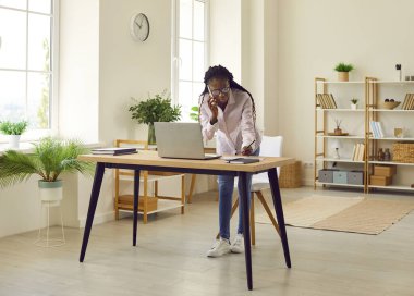 Kendine güvenen Afro-Amerikan iş kadınının tam boy portresi modern ofisindeki iş yerinin masasında dizüstü bilgisayarla çalışıyor ve cep telefonuyla konuşuyor..