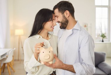 Mutlu genç çift, birbirlerine bakıyorlar ve kumbaraya para koyuyorlar. Gülümseyen adam ve kadın oturma odasında kucaklaşıyorlar, aile bütçesini birlikte planlıyorlar. Tasarruf ve ekonomi kavramı.