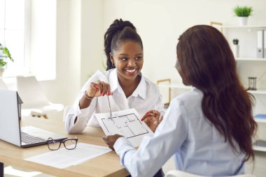 Gülümseyen emlakçı alıcıya ev planı sunuyor. Mutlu Afrikalı Amerikalı kadın emlakçı müşteriyle çalışır, tavsiye verir, konut planı gösterir ve tahmini fiyat hakkında konuşur.