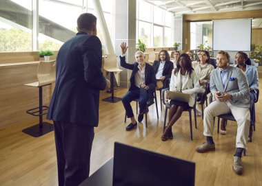 Gülümseyen olgun bir adam iş toplantısında soru sormak ya da cevap vermek için elini kaldırıyor. Bir grup şirket çalışanı ya da bir ekip sandalyede oturup konferansta liderlerini dinliyor..