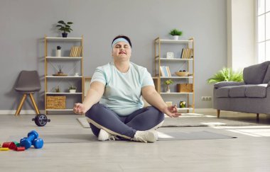 Evde yoga yapan aşırı kilolu gülümseyen bir kadın. Ayrıca spor giyim ve saç bandı giymiş, nilüfer pozisyonunda oturmuş meditasyon yapan boyunda bir kadın. Kilo verme egzersizi