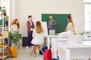 Ders ve öğretim. Beyaz tahtanın yardımıyla akıllı erkek öğretmen üniversite öğrencilerine eğitim materyallerini açıklayan dersler veriyor. Öğrenciler öğretmenin yanında durup dikkatle dinlerler..