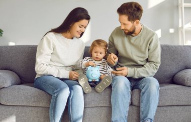 Gülümseyen ebeveynler bebeğe para biriktirmeyi öğretiyor. Anne, baba ve çocuk evdeki kanepede oturmuş domuz kumbarası tutuyorlar. Mutlu genç aile domuz kumbarasında para biriktiriyor.