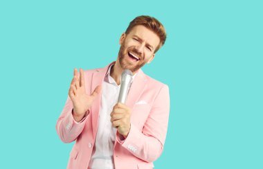 Mikrofonda şarkı söyleyen ve el kol hareketi yapan yakışıklı duygusal bir genç. Zarif pastel renkli pembe ceket giyen çekici sakallı adam açık mavi renkli stüdyo arka planında izole edilmiş performans sergiliyor.