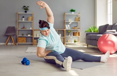 Kilolu, tombul bir kadın evde spor yapıyor. Yorgun, şişman, tombul, spor giyimli genç bir kız evde spor minderinde oturuyor ve yan eğme egzersizi yapıyor.