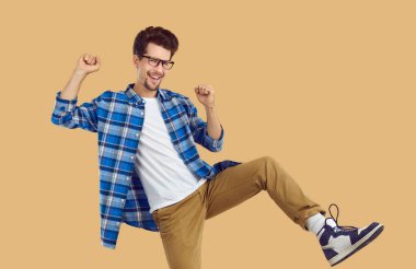 Olumlu duygular. Neşeli, funky hipster dans eden adam stüdyodaki bej arka planda mutlulukla dans ediyor. Günlük gömlekli, tişörtlü, pantolonlu ve spor ayakkabılı komik sakallı adam dans ediyor, gülüyor ve el sallıyor.
