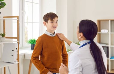 Kadın doktor, klinikte ya da hastanede çocuk hastasıyla konuşurken arkada oturuyor. Çocuk doktoru genç bir çocuğu muayene ediyor ve muayene odasında danışmanlık yapıyor..