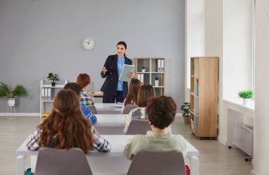 Ortaokuldaki kız arkadaş canlısı gülümseme öğretmeni. Okul çocukları ve sınıf arkadaşları sıralarda oturuyor ve sınıfta çalışıyor. Eğitim, bilgi ve okul konseptine geri dönüş.