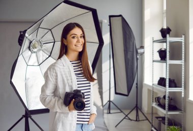 Fotoğraf stüdyosunda çalışan gülümseyen profesyonel kadın fotoğrafçı. DSR kameralı güzel genç bir kadının portresi. Stüdyoda duruyor. Profesyonel fotoğraf ekipmanıyla.
