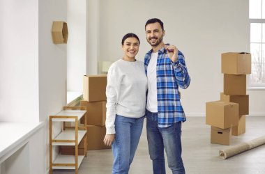 Mutlu aile çifti taşınma gününde yeni evin anahtarını elinde tutuyor. Gülümseyen eşler oturma odasında elinde karton kutularla kameranın anahtarlarını gösteriyorlar. Taşınma günü, emlak.