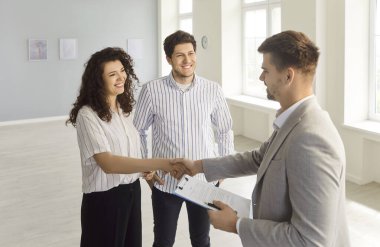 Mutlu gülümseyen genç müşteriler profesyonel bir emlakçıyla el sıkışıyor ve yeni dairelerinde mal satın alarak anlaşma yapıyorlar. Aile ev sahibi olmaya hazır. Emlak piyasası kavramı