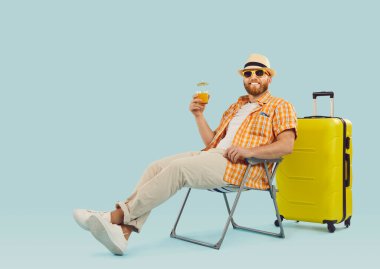 Mutlu gezgin yaz tatilinin tadını çıkarıyor. Tatil gömleği, güneş şapkası ve gözlüklü, gülümseyen sakallı adam kokteyl içiyor ve açık mavi fotokopi stüdyosunun arka planında sarı valizin yanında oturuyor.