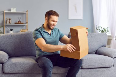 Evdeki kanepede karton kutuyu açan mutlu genç adam, yakın plan. Çevrimiçi sipariş teslimatı, online alışveriş, paketleri açma. Evinize mal sipariş etmek, neşe dolu alışverişkolik