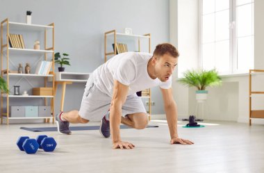 Beyaz tişörtlü yakışıklı genç adam evde spor egzersizleri yapıyor. Atletik adam oturma odasında yerde egzersiz yapıyor. Spor, spor ve ev eğitimi konsepti.