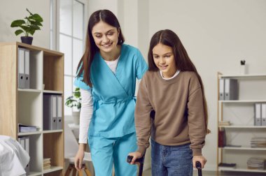 Genç ve neşeli bir esmer hemşirenin portresi çocuk bir kızın koltuk değnekleriyle yürümesine yardım ediyor. Dost canlısı fizyoterapist ya da çocuk doktoru engelli bir kızın rehabilitasyonuna yardım ediyor.