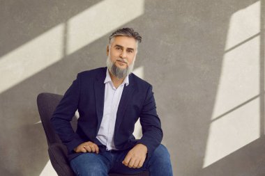 Ofiste poz veren orta yaşlı, sakallı, gri saçlı iş adamının portresi. Yakışıklı, kendine güveni tam bir iş adamı. Sandalyeye oturup kameraya bakıyor.