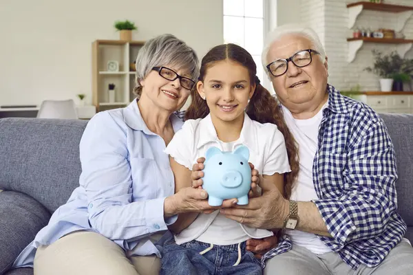 Şirin bir kız torun, büyük anne ve büyük babasıyla birlikte evdeki kanepede oturmuş elinde mavi domuz kumbarasıyla kameraya neşeli bir şekilde bakıyor. Para ve finansal okur yazarlık kavramı biriktiriliyor.