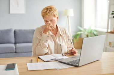 Uzun saatler çalıştıktan sonra baş ağrısı ya da göz yorgunluğu çeken yaşlı bir kadın iş yerinde dizüstü bilgisayarla ya da burun köprüsüne masaj yaparak masasında oturuyor..