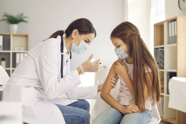 Female nurse giving an arm injection to a child. Little kid in a face mask getting a shot at a modern vaccination center. Doctor in white gloves gives a new effective Covid 19 vaccine to a little girl