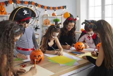 Arts and Crafts studio teacher doing creative activity with kids. Children dressed up as pirates, witches and vampires having fun Halloween party with their young English tutor at after school club