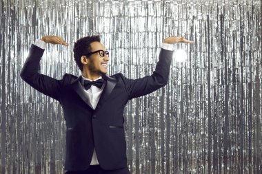 Young positive dark-skinned man in tuxedo performs funny dance moves on silver shiny background. Happy guy with funny expression looks away and dances with his hands up. Party concept. Banner.