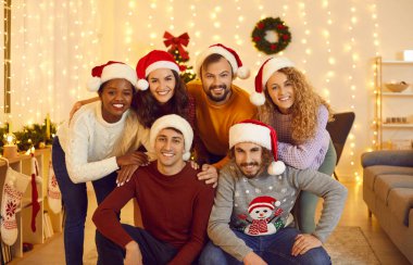 Portrait of cheerful young friends in Santa hats posing in a cozy living room with Christmas decor. Smiling multiracial international people gather at home enjoying the New Year celebration.
