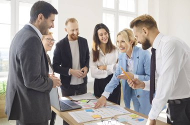 Group of smiling business professionals gathering around desk, analyzing financial indicators in diagrams and charts. Team of colleagues discussing sales performance, working in collaborative project.
