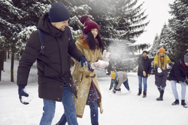 Young cheerful group of friends having fun together playing snowballs in snowy forest. Smiling man and woman dodge snowballs thrown at them by friends in background. Winter fun concept.
