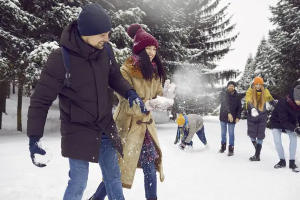 Young cheerful group of friends having fun together playing snowballs in snowy forest. Smiling man and woman dodge snowballs thrown at them by friends in background. Winter fun concept.