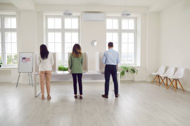 Backside view of diverse group of citizens is seen polling on USA election day. The bustling scene unfolds in a voting center, where individuals from all walks of life gather to cast votes in booths.