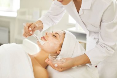 Kozmetik uzmanı, spa salonundaki genç, rahat, gülümseyen bir kadına nemlendirici maske takıyor. Güzellik kliniğindeki güzellik uzmanından güzellik prosedürü alan bir kız müşterinin portresini yakından çek