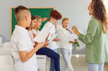 Portrait of a young school children standing with their young female teacher having music lesson in the classroom. Kids singing in a choir holding white sheets preparing for performance.
