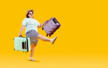 Happy funny woman going on summer vacation. Cheerful fat overweight woman in T shirt, shorts, sun hat and sunglasses holding green and purple travel suitcases and walking isolated on yellow background