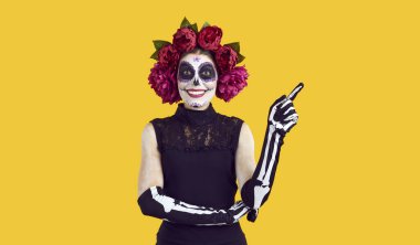 Happy lady with sugar skull Catrina makeup and wreath of pink roses on head smiles and points finger up. Woman dressed in costume on Halloween or Day of the Dead standing isolated on yellow background