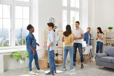 Group of diverse friends meeting and hanging out together. Young multiracial mixed race people in casual clothes standing in room at home, talking to each other, discussing latest events