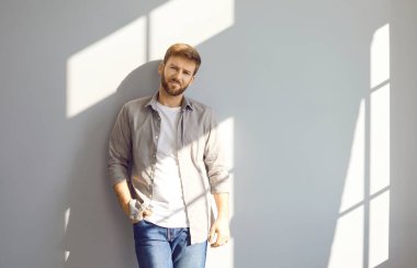 Portrait of handsome man in casual clothes. Young guy with short beard and mustache in grey shirt, white T shirt and blue jeans standing with hand in pocket against light grey wall. Fashion concept