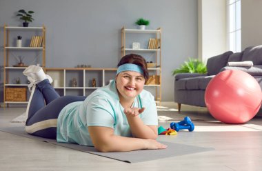 Portrait of a funny cheerful young fat woman wearing sportswear lying on the floor on yoga mat at home and looking at the camera. Workout, sport exercises, fitness and body positive concept.